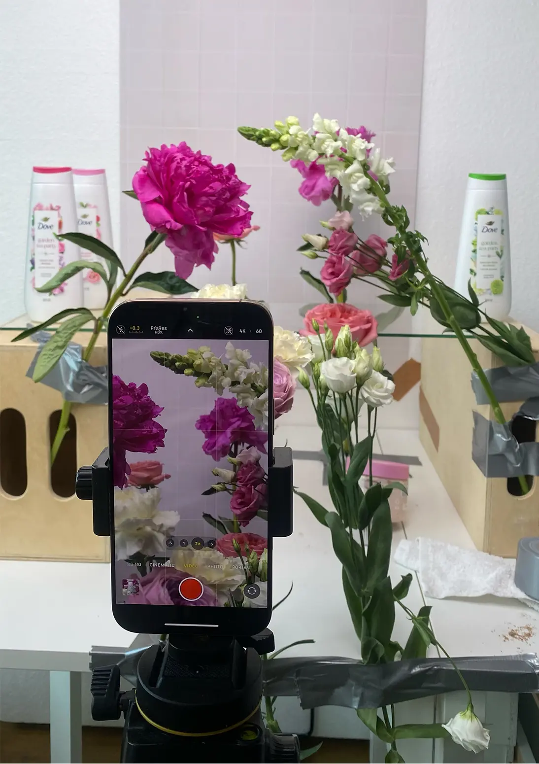 Smartphone on a tripod filming a floral product setup on a table, with flowers arranged around packaged toiletries, showing a low-fi, in-progress filming setup.